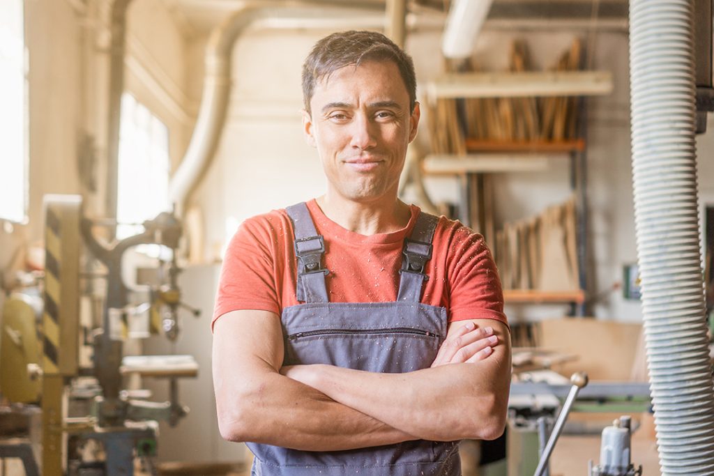 Carpenter in uniform standing in professional light workshop Content male woodworker in overall looking at camera with crossed arms while standing in professional light carpentry with various equipment